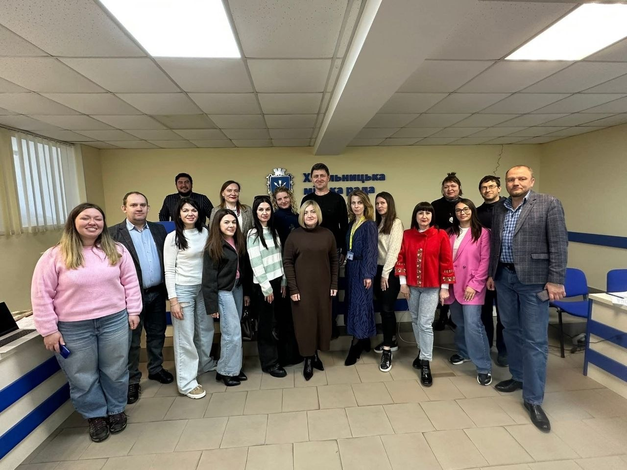 Група людей позує для спільного фото в офіційному приміщенні на фоні стіни з написом «Хмельницька міська рада» та гербом міста. Учасники зустрічі стоять у кілька рядів у залі з офісними меблями.