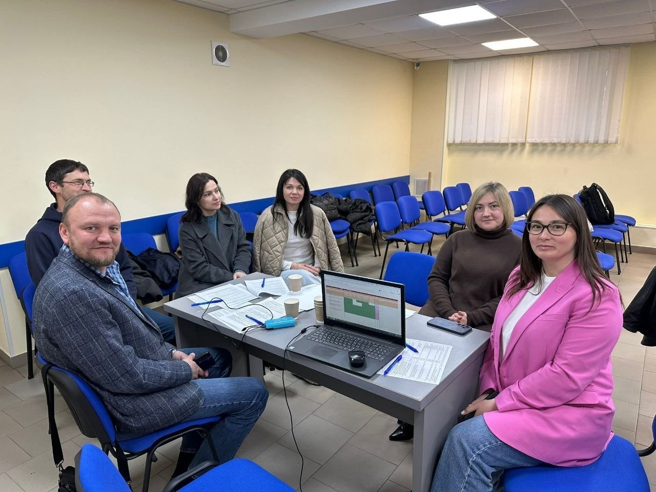 Група людей проводить робочу зустріч у залі з синіми стільцями. Учасники сидять за столом, на якому лежать документи, паперові стаканчики та відкритий ноутбук із робочою таблицею на екрані.