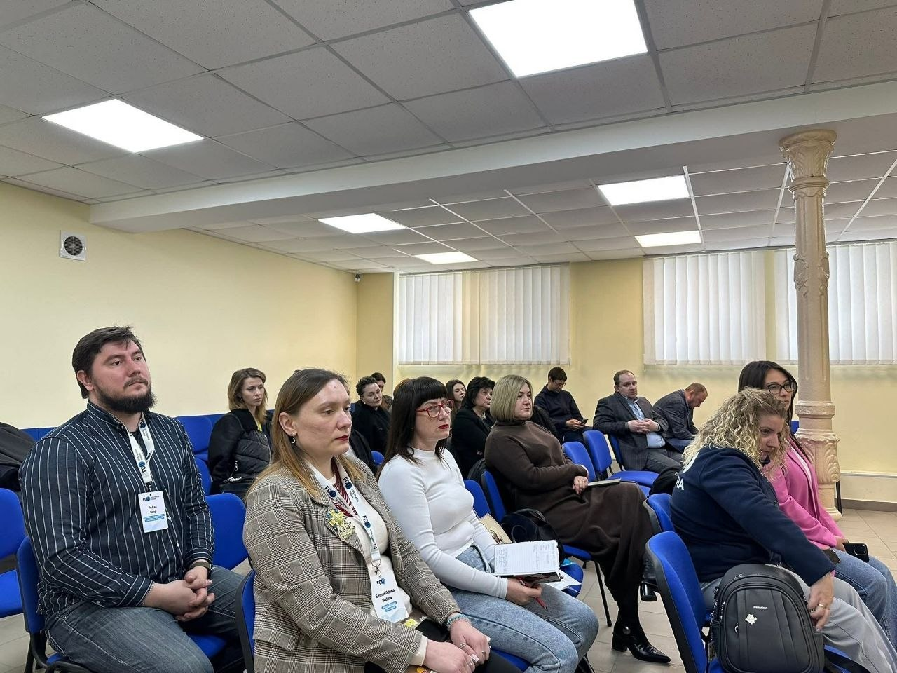 Група людей сидить у залі на синіх стільцях під час офіційного заходу або тренінгу. Учасники на передньому плані мають бейджі на шиї та зосереджено слухають спікера. Приміщення освітлене стельовими лампами, на вікнах — жалюзі.