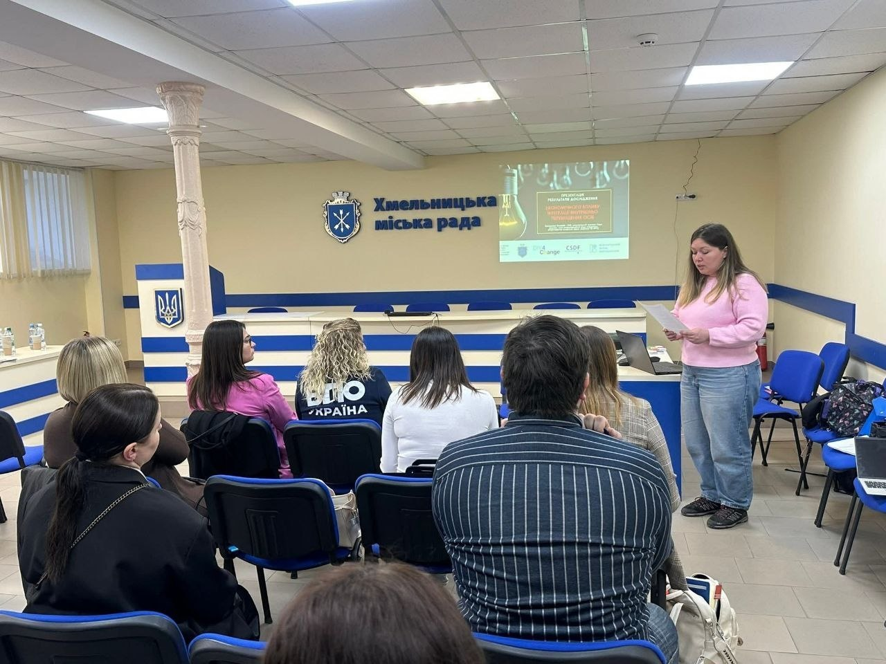 Жінка у рожевому светрі виступає перед аудиторією в залі Хмельницької міської ради. На стіні за нею — герб міста, напис «Хмельницька міська рада» та проєкція презентації на екрані. Слухачі сидять у кріслах спиною до камери.