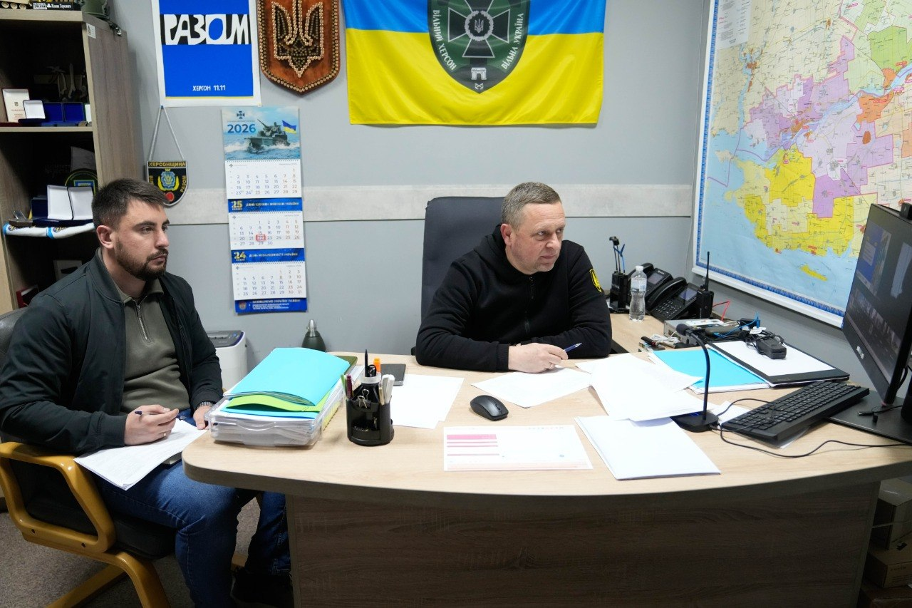 Двоє чоловіків проводять робочу зустріч у кабінеті. Один сидить за столом із документами та комп'ютером, інший — поруч із блокнотом. На стіні за ними висять прапор України з емблемою, мапа області, календар на 2026 рік та державна символіка.