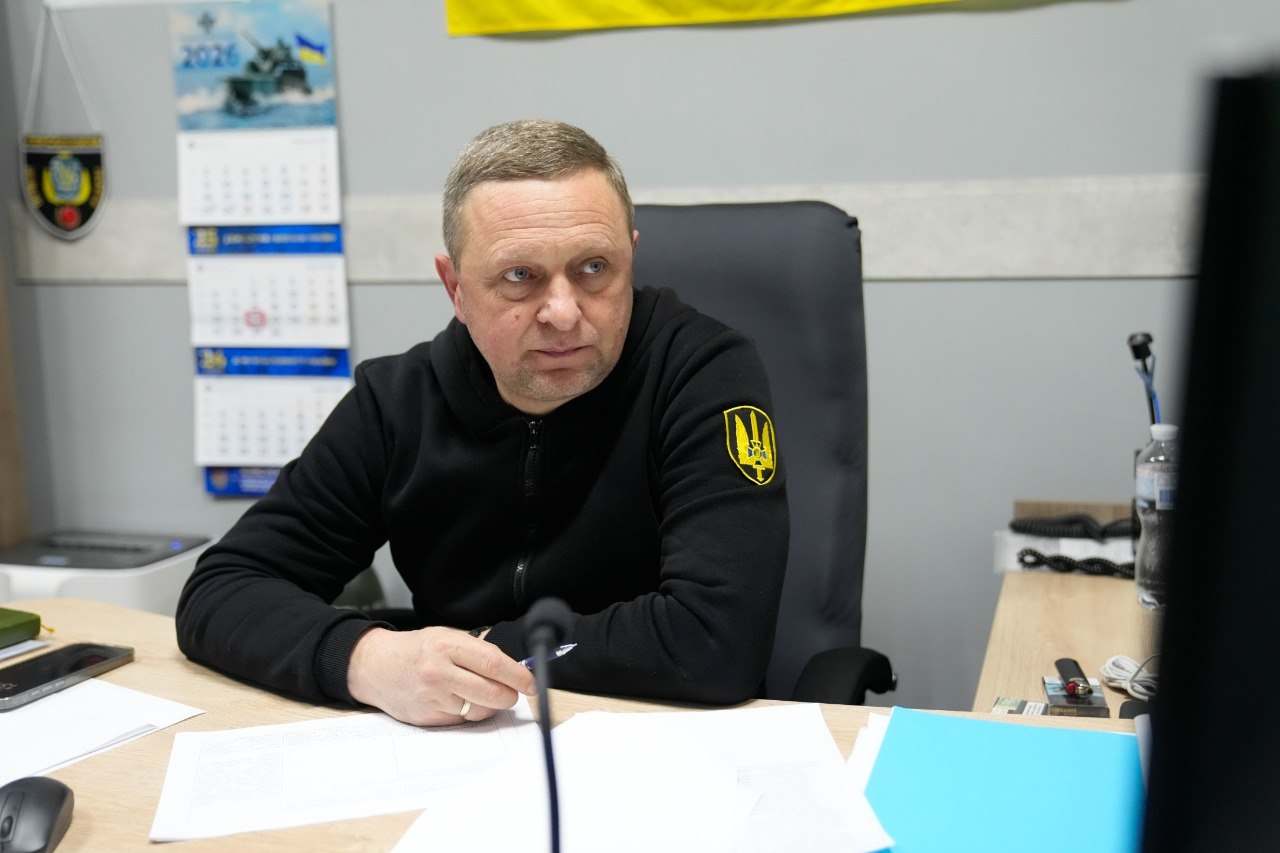 Чоловік у чорному худі з гербом України на рукаві сидить за робочим столом. Перед ним лежать папери та ручка, а на задньому плані видно календар на 2026 рік із зображенням військового корабля та синьо-жовтий прапор.