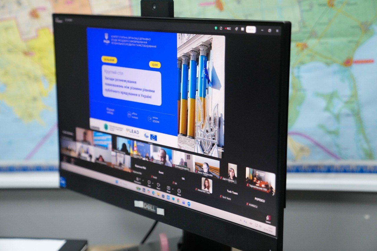 Монітор комп'ютера, на якому відображається онлайн-конференція в Zoom. На екрані презентація круглого столу Комітету Верховної Ради України, присвяченого розмежуванню повноважень між рівнями публічного врядування. У нижній частині — мініатюри вікон з учасниками відеозустрічі.