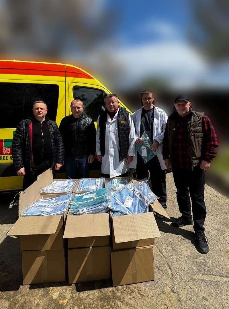 П'ятеро чоловіків, серед яких медики в білих халатах, позують поруч із відкритими коробками з медичним одягом. На задньому плані — яскраво-жовтий автомобіль швидкої допомоги та розмите зображення дерев.