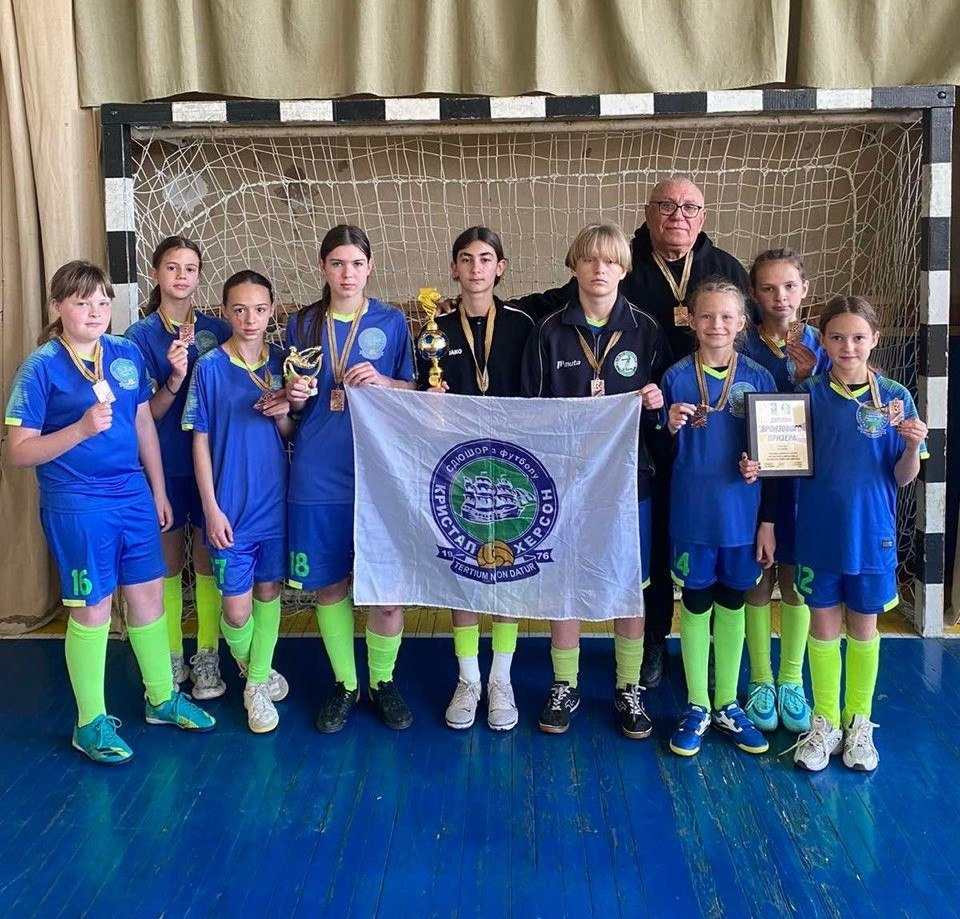 Дитяча футбольна команда дівчат у синьо-салатовій формі позує для фото на тлі футбольних воріт у спортзалі. Юні спортсменки з медалями та кубком тримають білий прапор із написом «СДЮШОР з футболу 