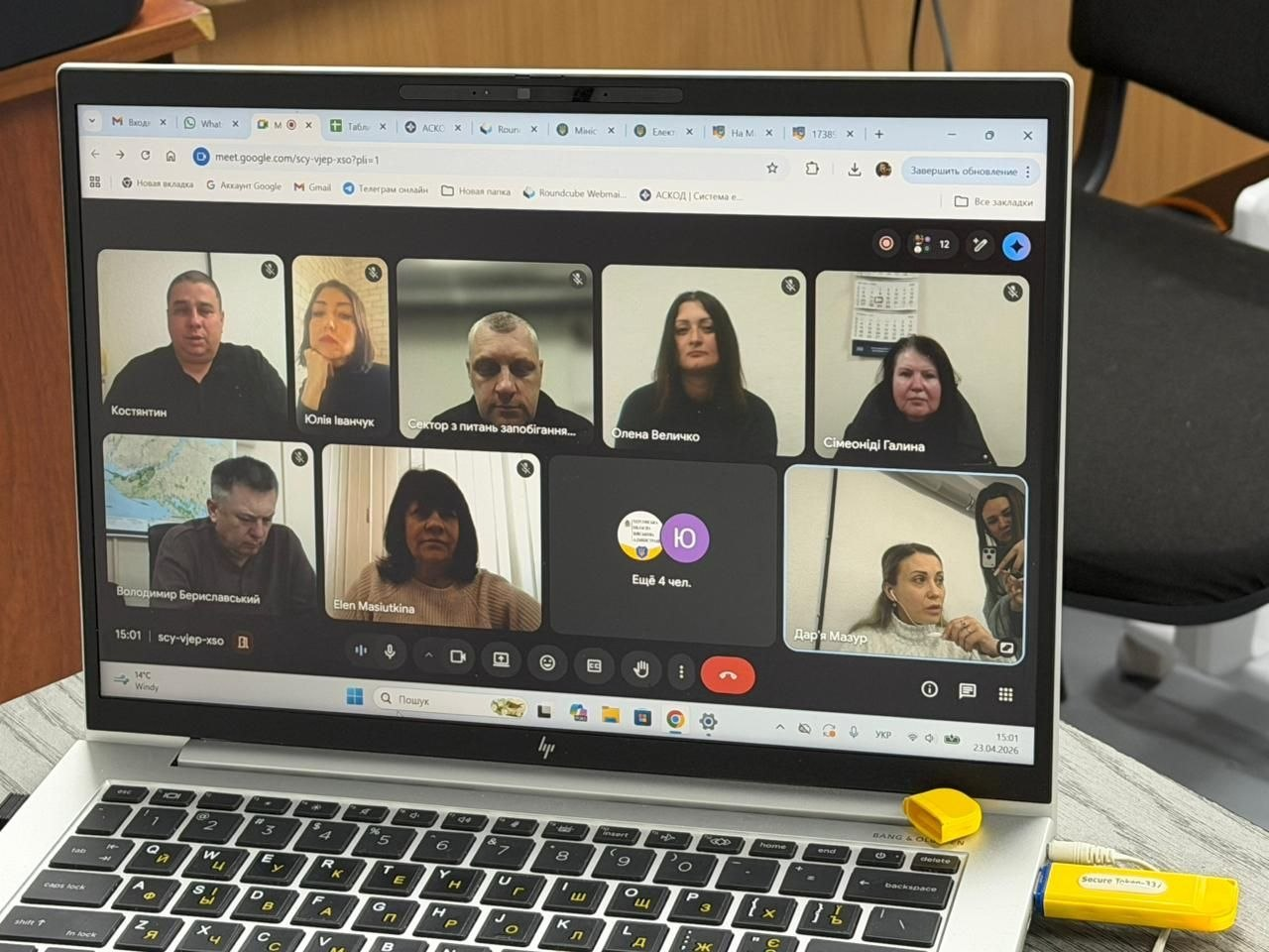 Ноутбук, на екрані якого відображається відеозустріч Google Meet із багатьма учасниками. У USB-порт комп'ютера вставлено яскраво-жовтий апаратний ключ безпеки.