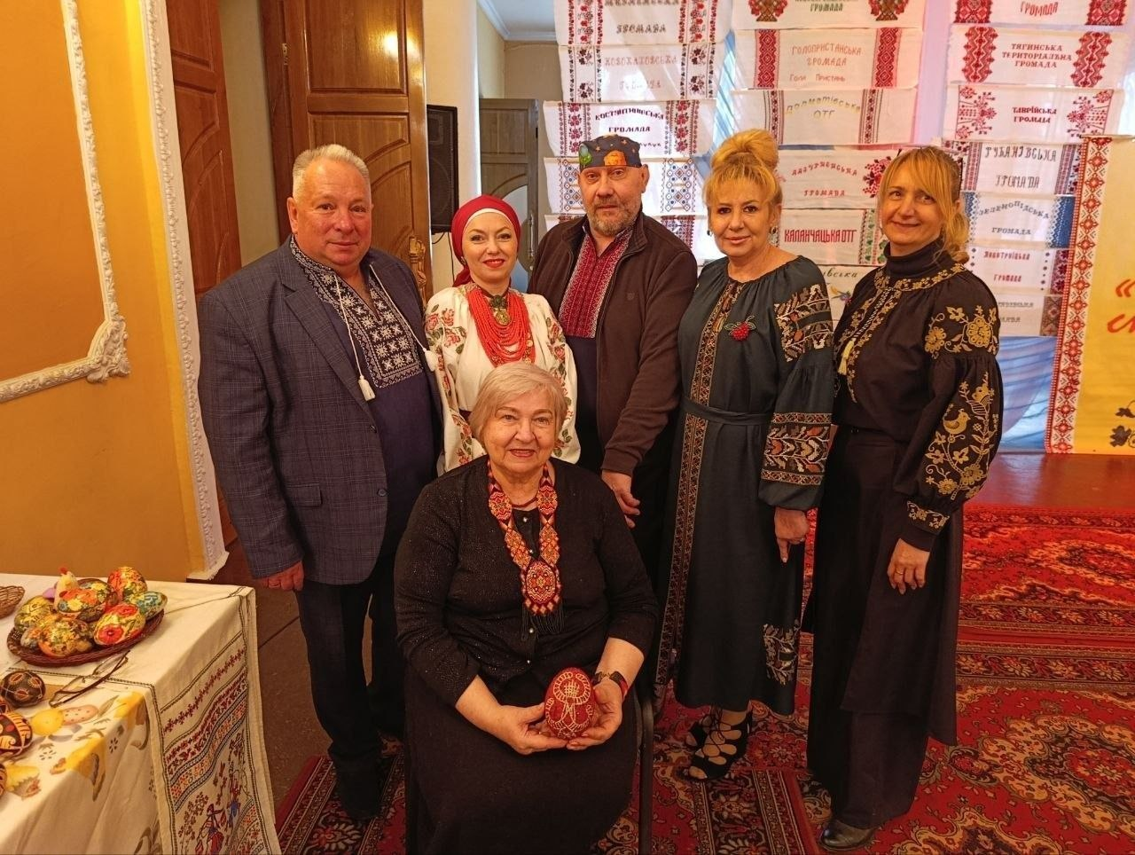 Група людей у традиційному українському та сучасному одязі з елементами вишивки позує на фоні вишитих рушників. Попереду сидить жінка з писанкою в руках, а збоку стоїть стіл із великодніми прикрасами.