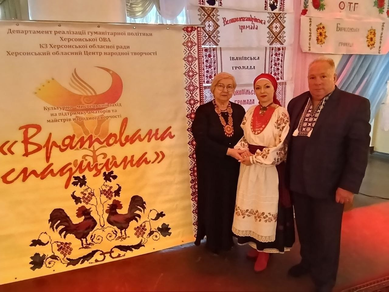 Три особи стоять біля великого банера культурно-мистецького заходу «Врятована спадщина». Жінка в центрі вдягнена у традиційний український стрій, двоє інших людей — у класичному одязі з елементами вишивки. На фоні видно вишиті рушники з назвами громад Херсонщини.