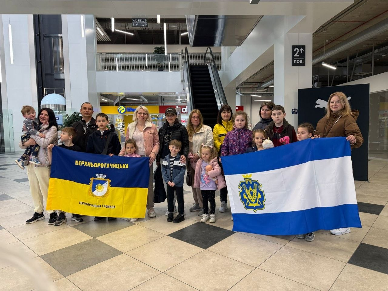 Група дорослих і дітей позує в торговому центрі, тримаючи два прапори: ліворуч — жовто-блакитний прапор Верхньорогачицької громади, праворуч — біло-синій прапор Херсонської області.