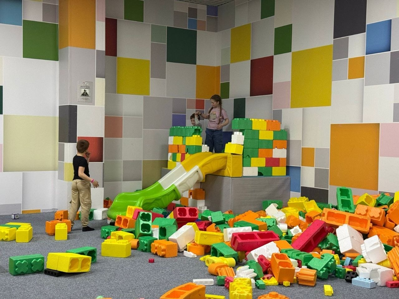 Діти граються в ігровій кімнаті з великим різноколірним конструктором. Вони побудували споруду з гіркою на фоні стіни, декорованої яскравими квадратами.