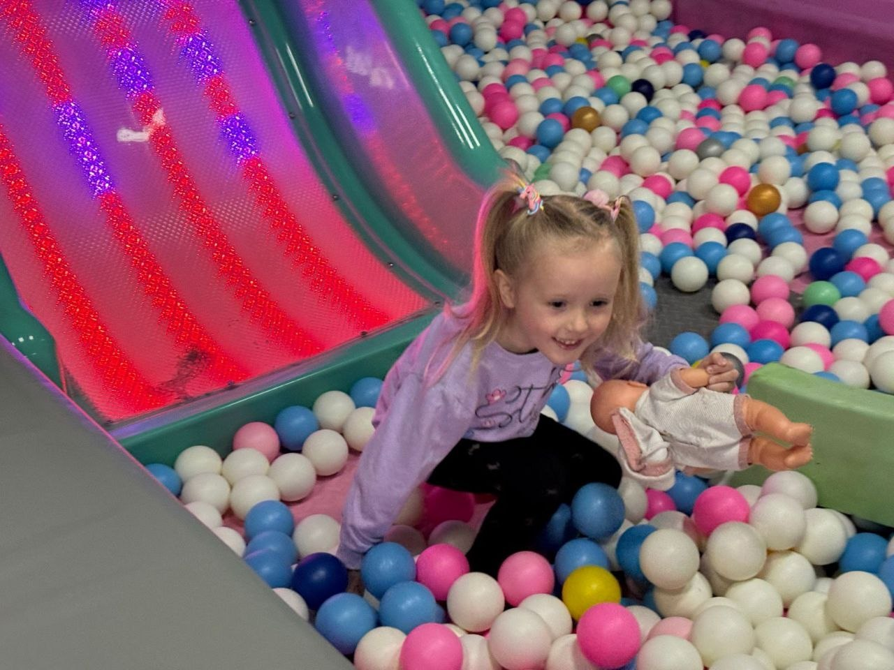 Дівчинка з лялькою в руках весело грається в сухому басейні з різноколірними кульками. Поруч розташована дитяча гірка з яскравим підсвічуванням.