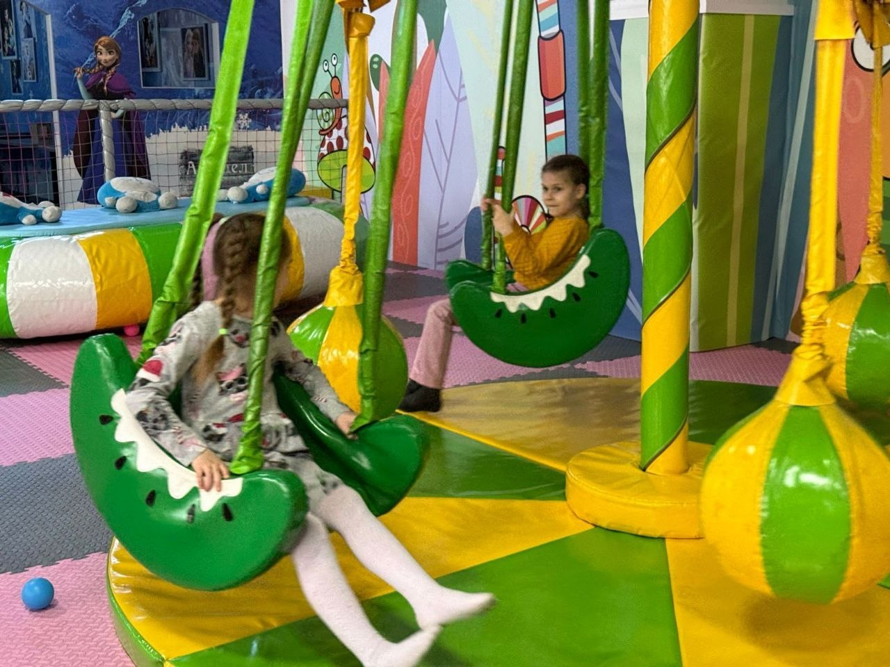 Двоє дітей катаються на дитячій каруселі з підвісними сидіннями у формі зелених часточок кавуна. Ігровий майданчик оформлений у яскравих зелених та жовтих кольорах.