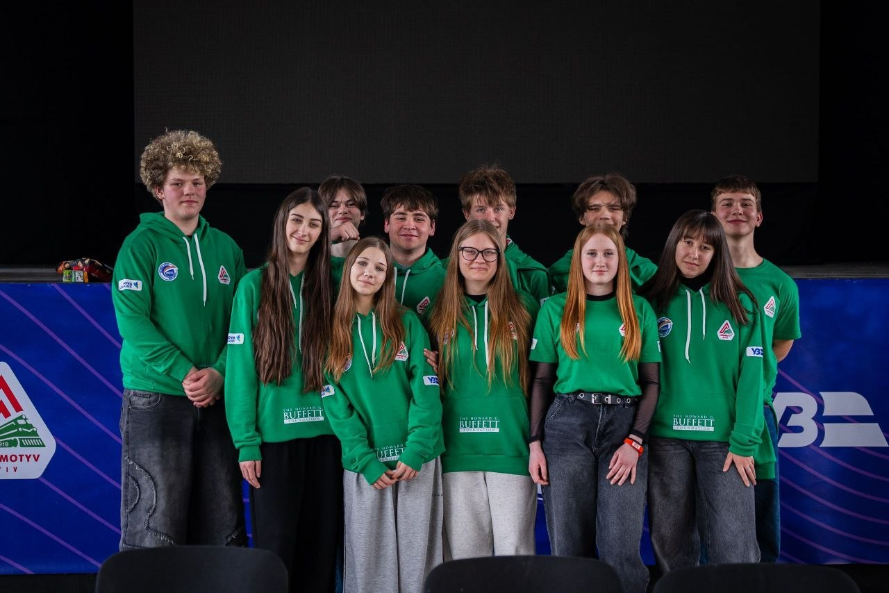 Група підлітків у зелених худі та футболках із логотипами позує на фоні сцени. Вони стоять у два ряди перед синім банером у приміщенні, що нагадує кінозал або конференц-хол.