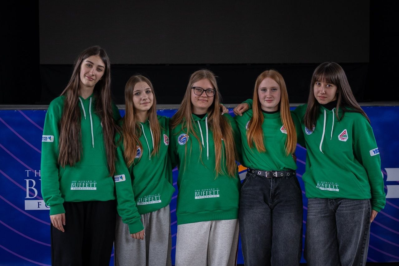 П’ять дівчат у зелених худі та футболках із логотипами фундації Говарда Баффета стоять у ряд на фоні сцени. Вони позують перед синім брендованим банером у темному залі.