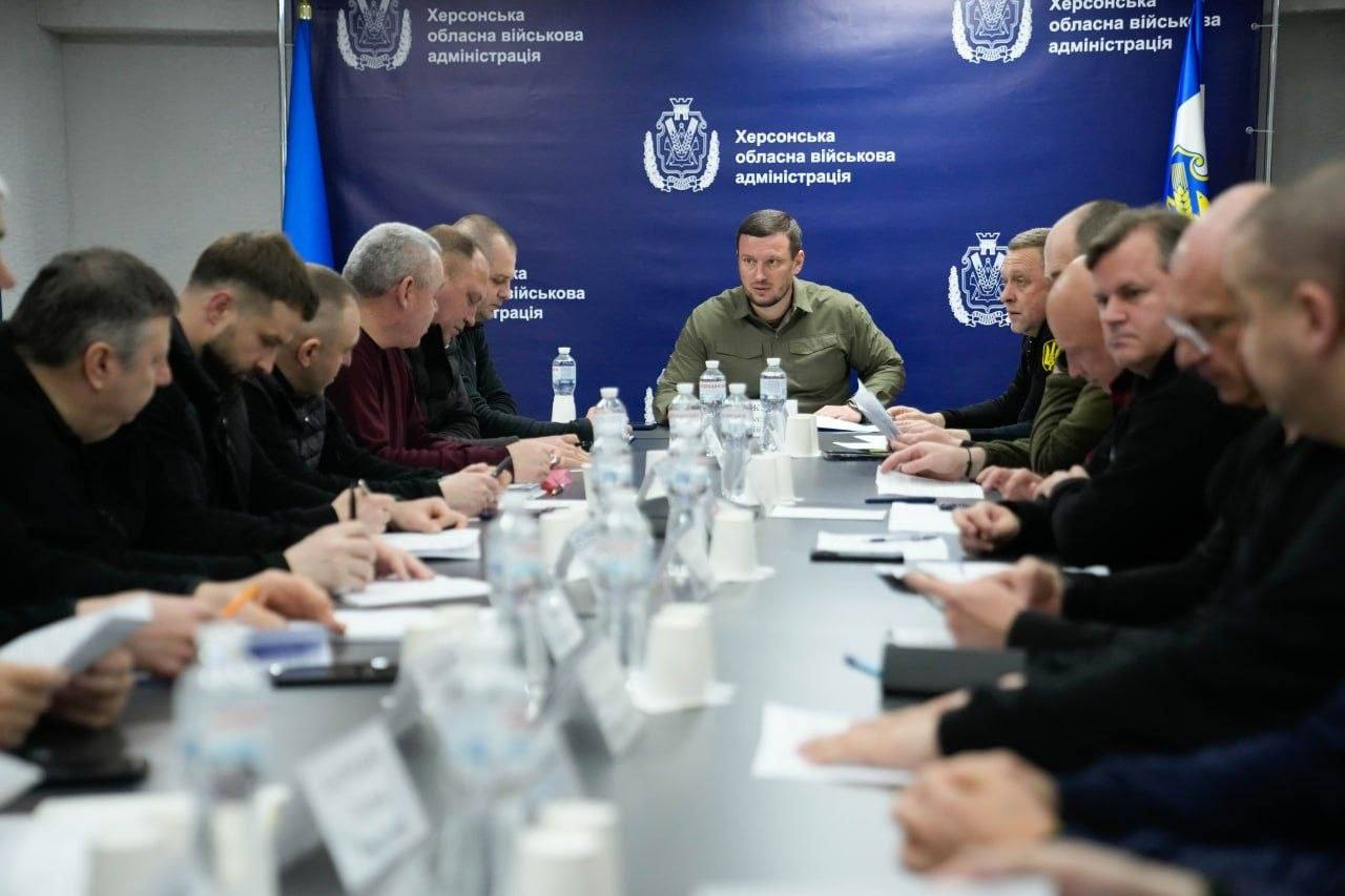 Група чоловіків проводить офіційну нараду за довгим столом. У центрі на фоні синього банера з офіційною символікою сидить керівник у мілітарі-одязі, а навколо нього — учасники зустрічі, які занотовують інформацію.