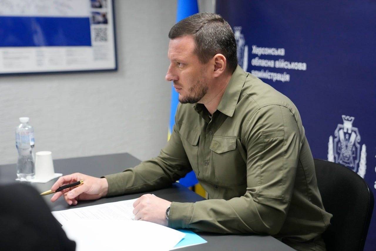 Чоловік у сорочці кольору хакі сидить за робочим столом у профілі. Він зосереджено дивиться перед собою, тримаючи ручку над паперами. На фоні видно частину синього банера з офіційною символікою та державний прапор.