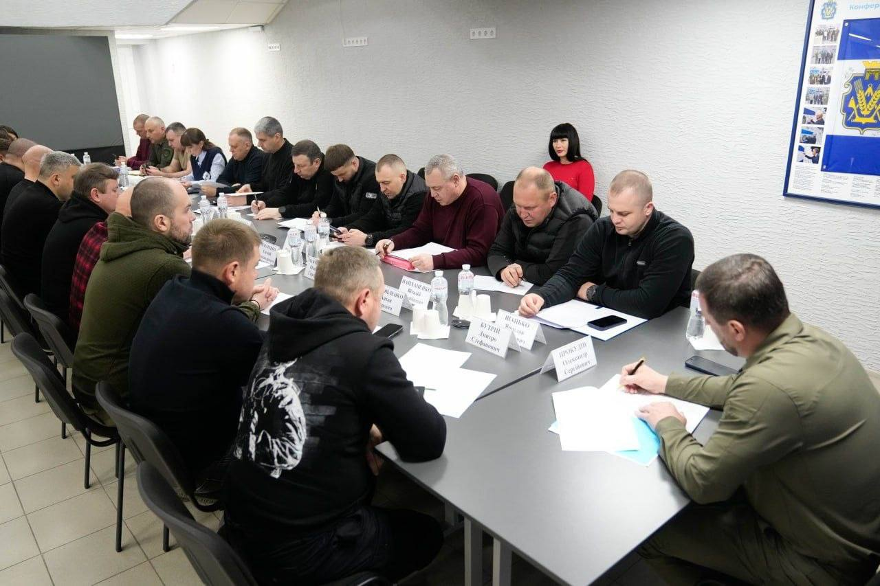 Велика група людей сидить за довгим Т-подібним столом під час наради. Учасники зустрічі зосереджено працюють із документами та роблять записи. На столі перед ними стоять таблички з іменами та пляшки з водою.