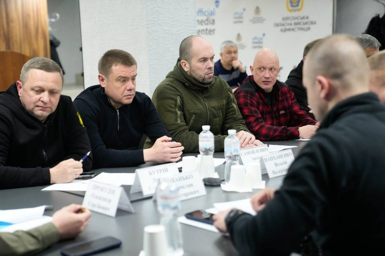 Четверо чоловіків сидять за столом під час офіційної зустрічі та уважно слухають доповідача. Перед ними стоять таблички з іменами та пляшки з водою. На фоні видно стіну з логотипами медіацентру та адміністрації.