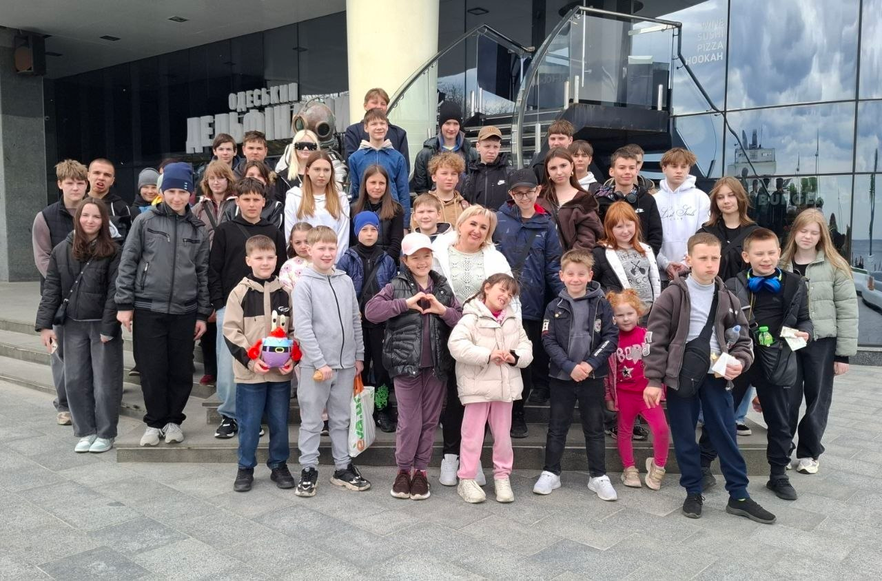 Велика група дітей та підлітків разом із дорослою жінкою стоять на сходах біля входу до сучасної скляної будівлі з написом «Одеський».