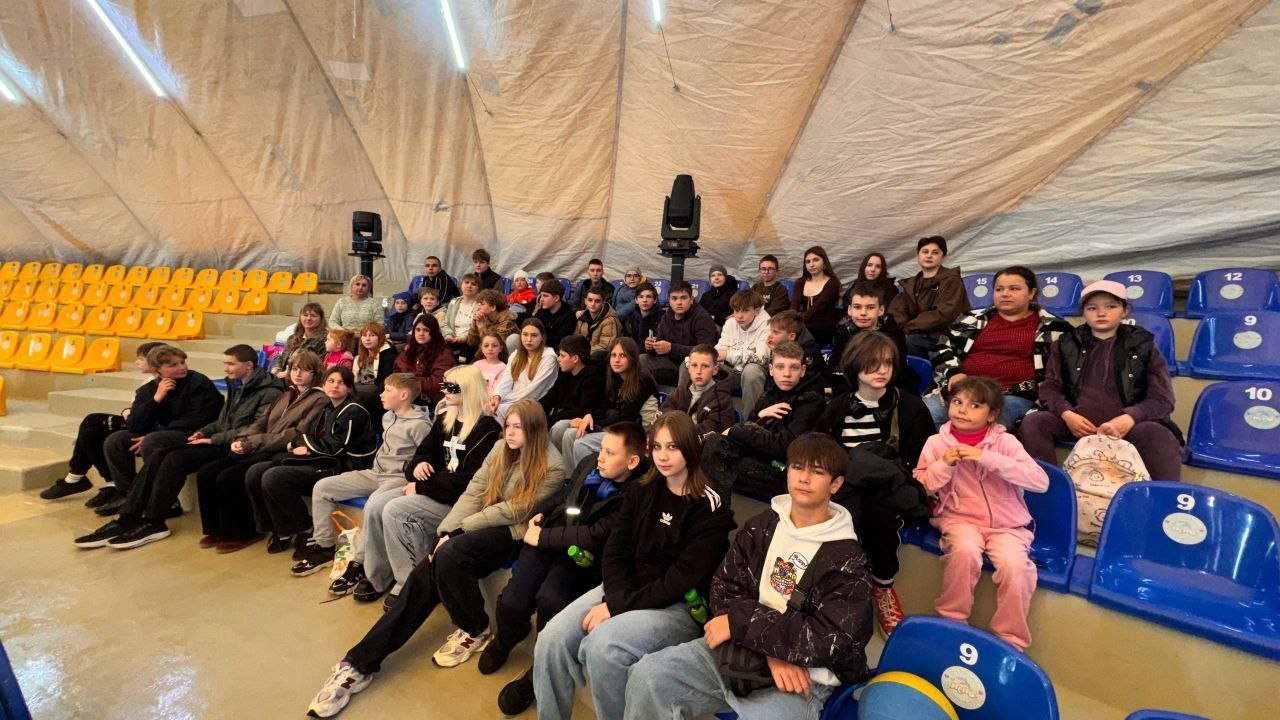 Група дітей та підлітків сидить на трибунах із синіми та жовтими кріслами всередині великої купольної споруди з тентовим дахом.