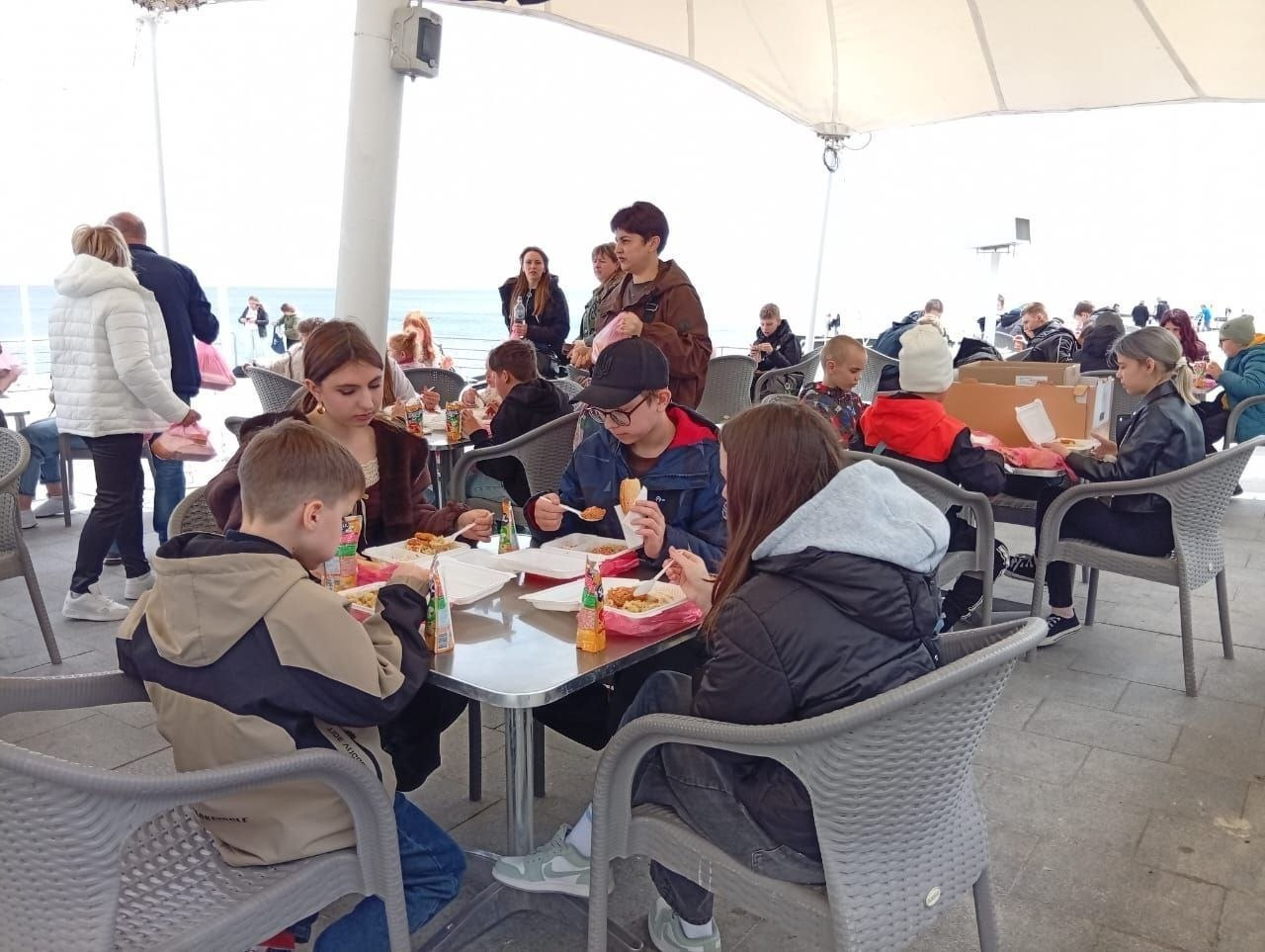 Група дітей та підлітків обідає за столиками у літньому кафе під білим тентом на фоні морського узбережжя.