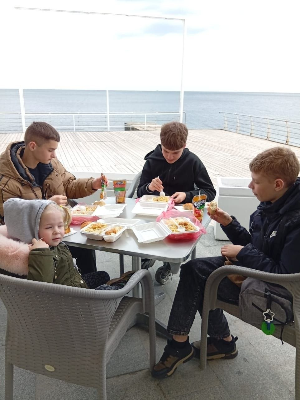 Четверо дітей та підлітків обідають за столиком на відкритій терасі біля моря. На столі стоять контейнери з їжею та пакетовані соки.