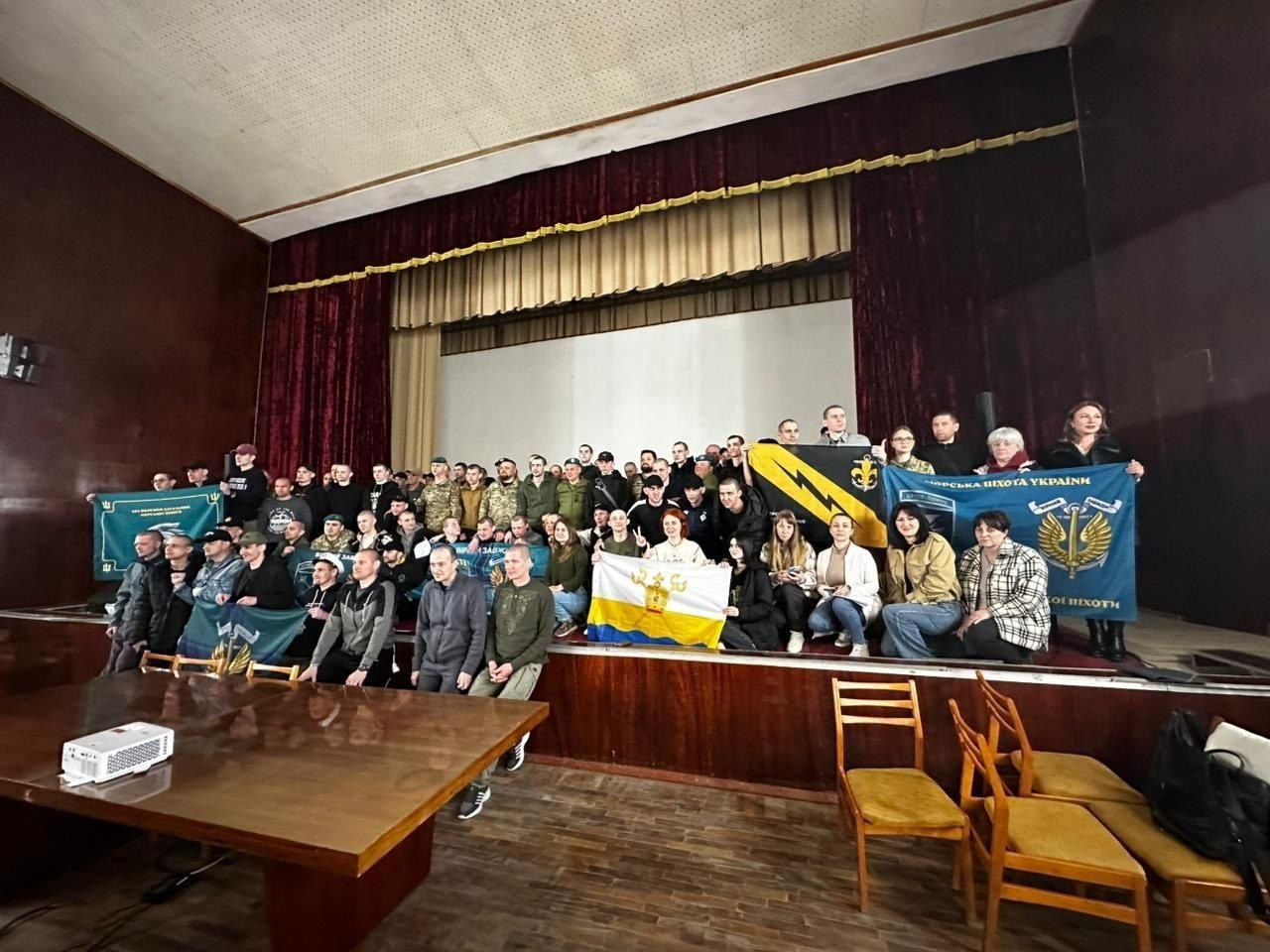 Велика група людей у цивільному та військовому одязі позує для спільного фото на сцені актового залу. Учасники тримають прапори з українською символікою та емблемами підрозділів. Перед ними стоїть великий дерев'яний стіл, а на передньому плані видно порожні стільці.