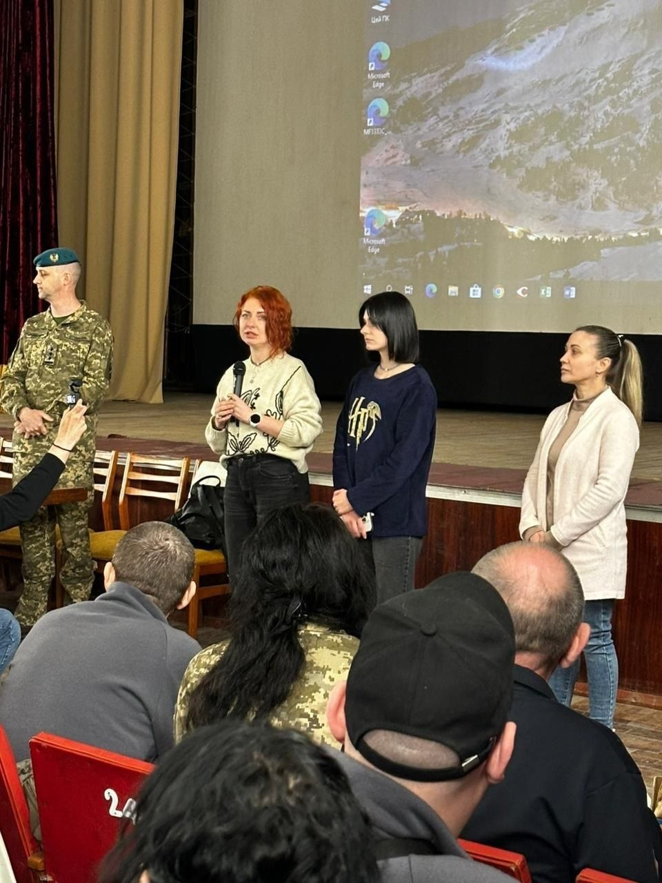 Група людей виступає на сцені перед аудиторією в актовому залі. Серед промовців — жінка з мікрофоном, двоє інших жінок та чоловік у військовій формі. На задньому плані видно великий екран із проекцією робочого столу комп'ютера.