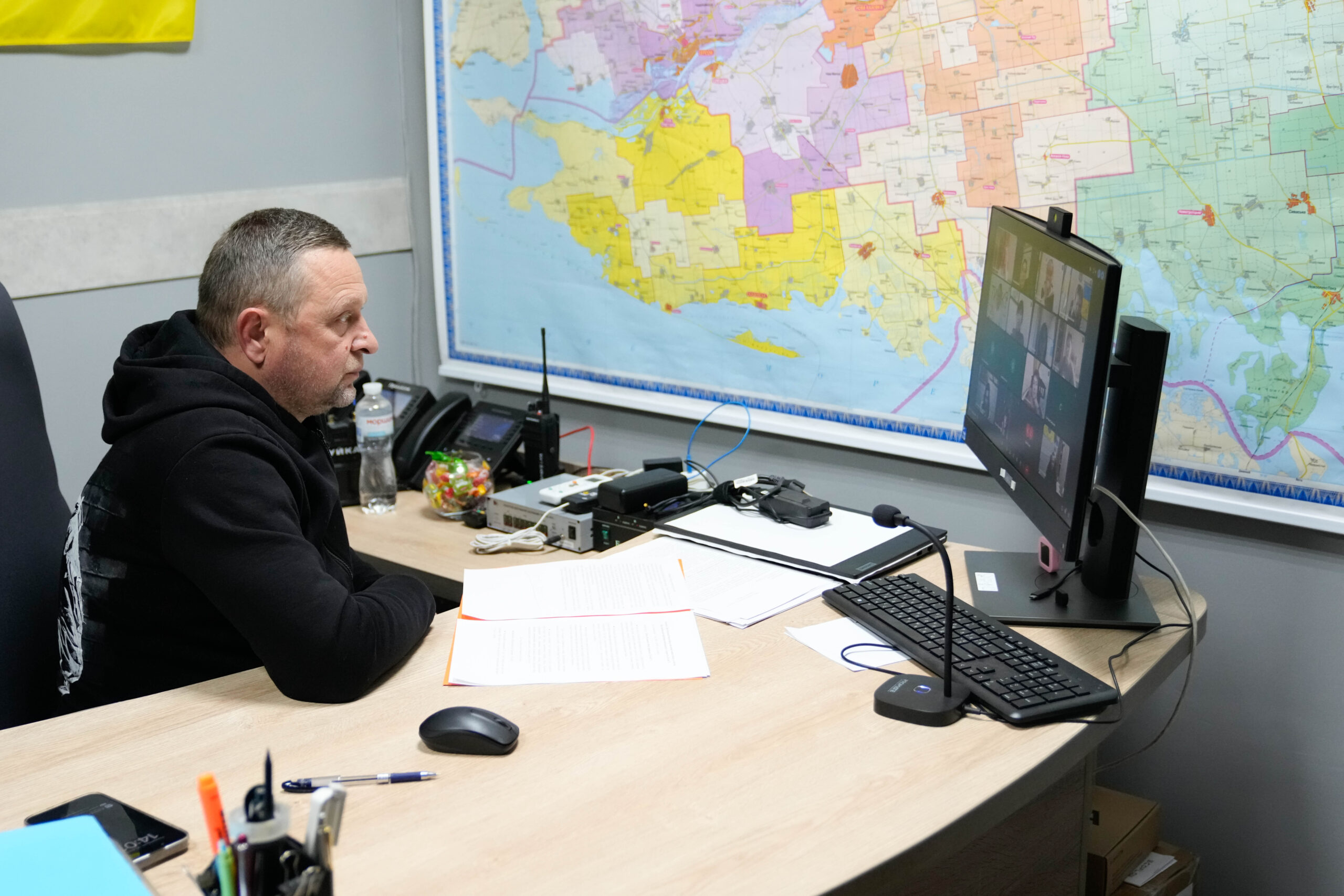 Чоловік за робочим столом бере участь у відеоконференції. Перед ним монітор, мікрофон та документи. На стіні позаду розміщена велика карта області.