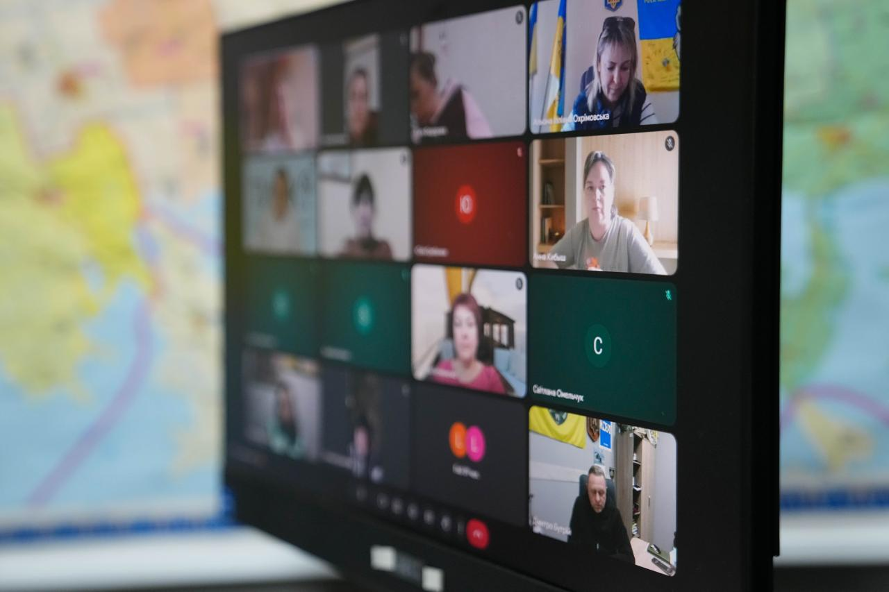 Монітор комп'ютера великим планом, на якому відображено інтерфейс відеоконференції з багатьма учасниками. Екран розташований на фоні розмитої настінної карти області.