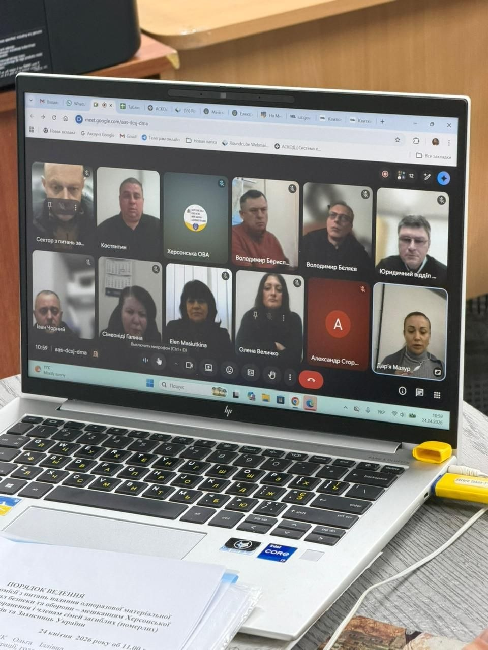 Ноутбук великим планом, на екрані якого відкрито відеоконференцію Google Meet із багатьма учасниками. Перед ноутбуком лежить документ із заголовком «Порядок ведення», а в роз’єм вставлено яскраво-жовтий USB-накопичувач.