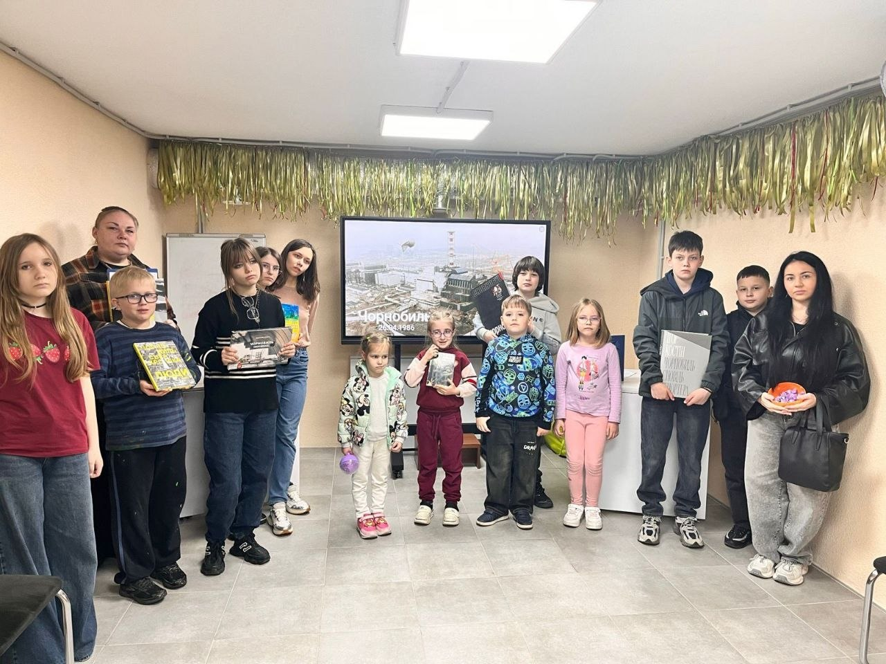Група дітей та молоді різного віку стоїть у світлій кімнаті перед великим екраном, на якому зображено Чорнобильську АЕС. Дехто тримає в руках книги або плакати. Приміщення прикрашене золотистим дощиком під стелею.