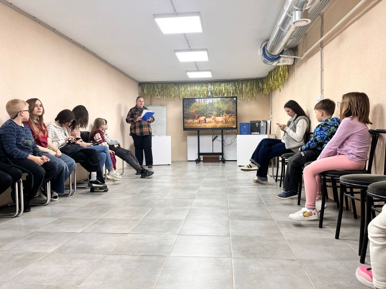 Група дітей та молоді сидить на стільцях вздовж стін у світлому приміщенні, схожому на навчальний клас або укриття. У центрі кімнати перед екраном стоїть доросла особа з папкою в руках і проводить презентацію. На великому моніторі відображено фото диких коней у природному середовищі.