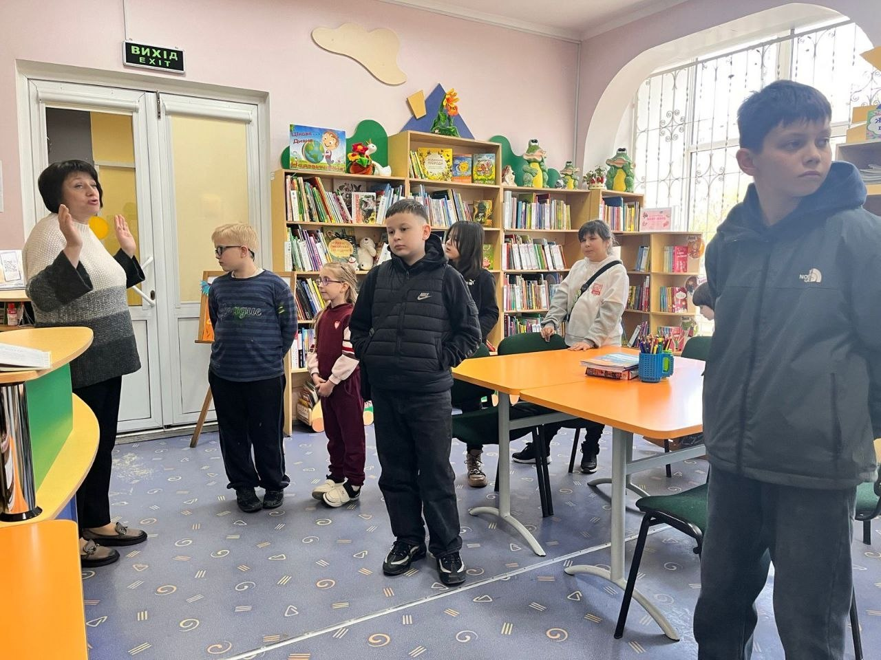 Група дітей стоїть у затишній бібліотеці або навчальній кімнаті. Доросла жінка зліва щось пояснює, жестикулюючи руками. На фоні видно стелажі з книгами та яскравий помаранчевий стіл.