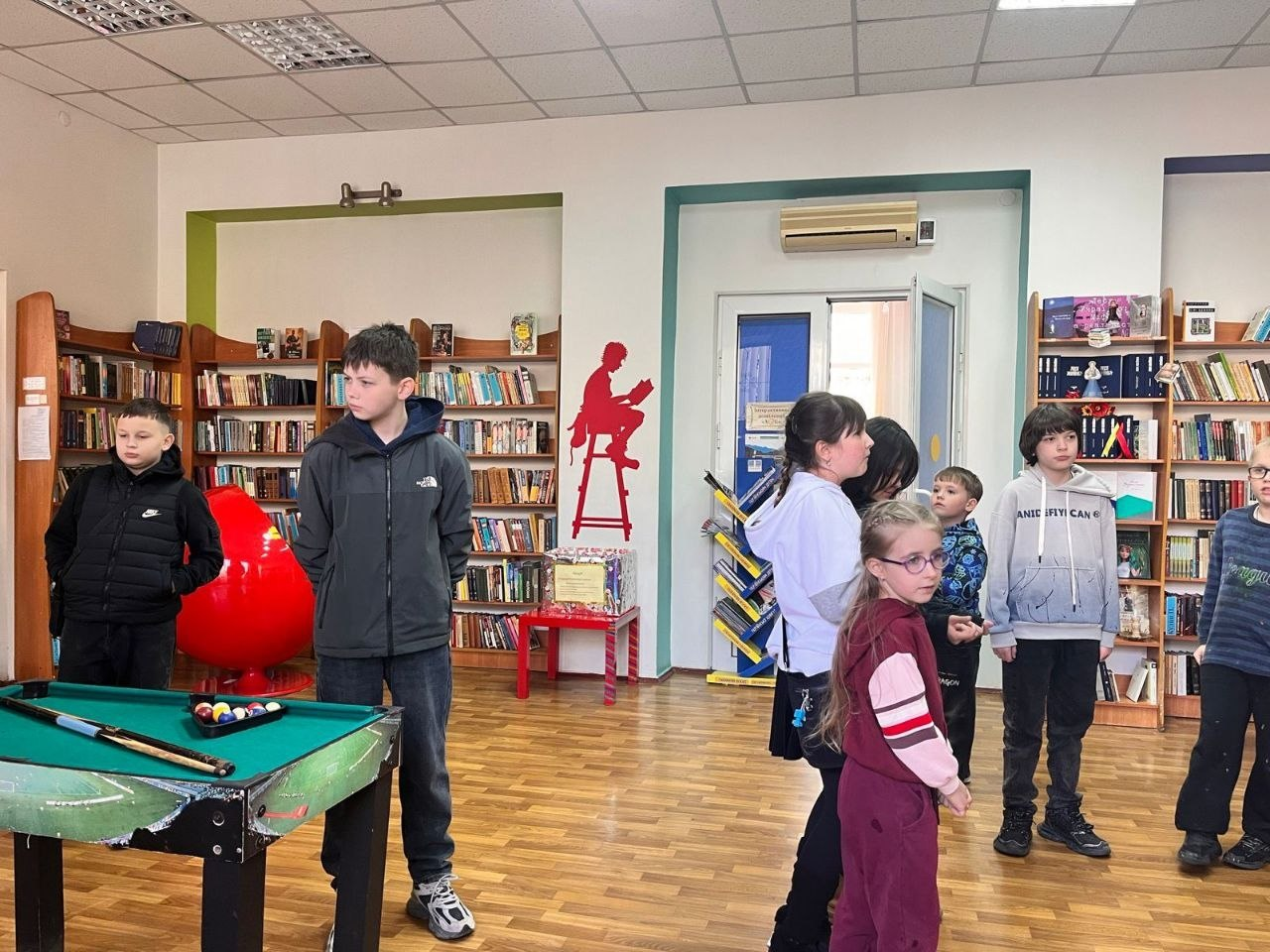 Група дітей у просторому світлому приміщенні бібліотеки або ігрової зони. На фоні видно стелажі з книгами та червоний декор на стіні, а на передньому плані зліва — невеликий більярдний стіл.