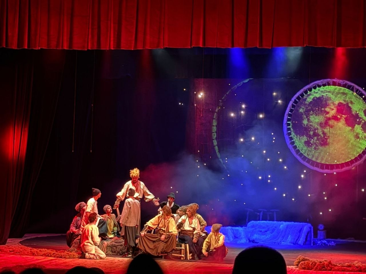 Театральна сцена з акторами у традиційному українському вбранні на фоні декорацій нічного неба з великим яскравим місяцем та зорями. Сцена освітлена синіми та червоними прожекторами, створюючи атмосферу містичності.