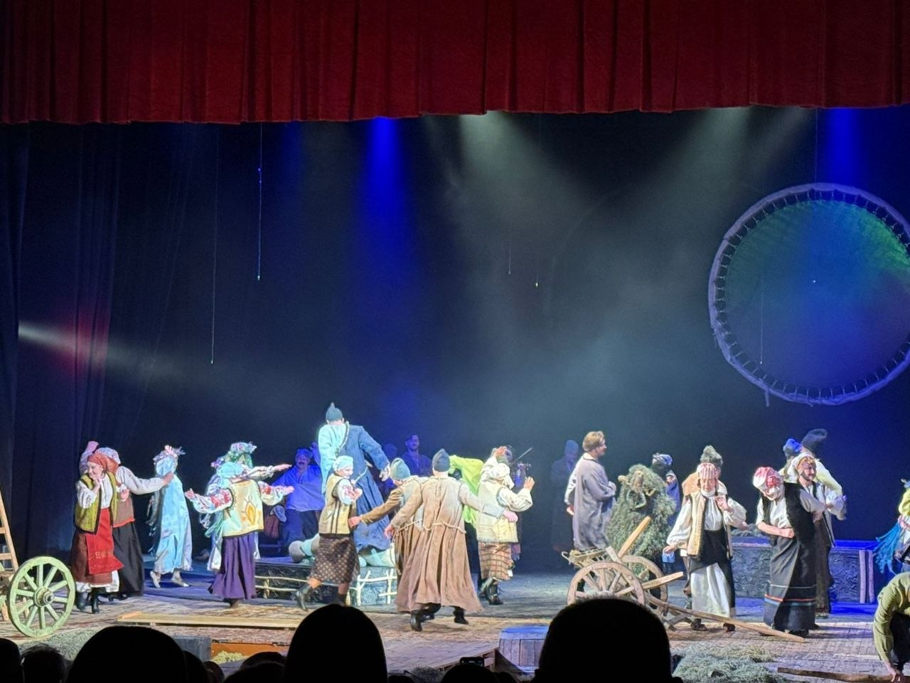 Театральна сцена, на якій група акторів у традиційних українських строях відтворює масову народну сцену. Декорації включають дерев’яне колесо та віз, а на задньому плані — велике кругле полотно. Сцена освітлена синіми та білими прожекторами.