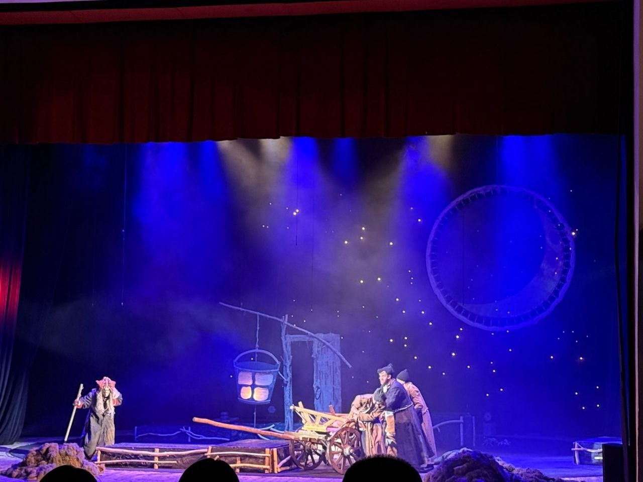 Театральна сцена у синьому освітленні, що імітує ніч. Актори в традиційному вбранні стоять біля дерев’яного воза та криниці-журавля з відром. На задньому плані — декорації зоряного неба та великого кола.