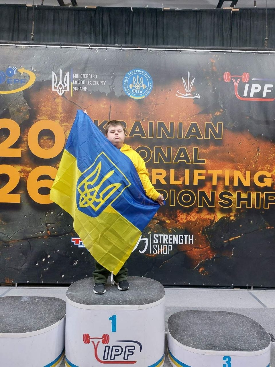 Юний спортсмен у жовтому худі стоїть на першій сходинці п'єдесталу пошани, тримаючи державний прапор України. Позаду нього — банер змагань з пауерліфтингу «Ukrainian National Powerlifting Championship 2026» із логотипами Міністерства молоді та спорту України та IPF.