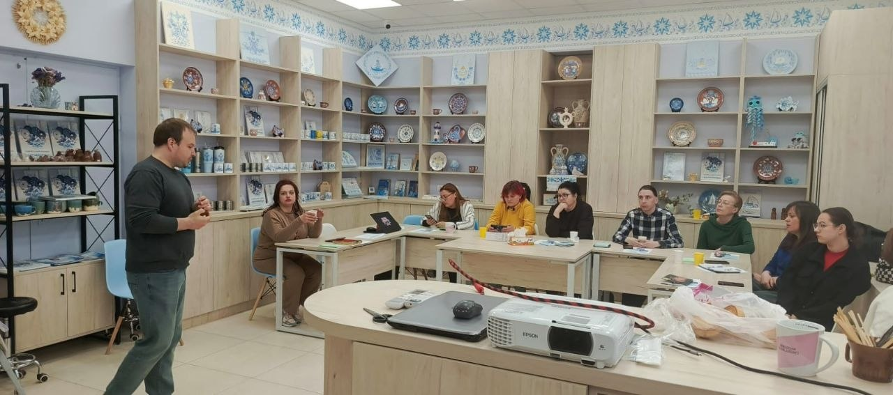 Група людей під час робочої зустрічі або майстер-класу у світлому приміщенні. Один чоловік стоїть і говорить, інші сидять за столами та слухають. Кімната оздоблена численними полицями з декоративним керамічним посудом та виробами з народним розписом.