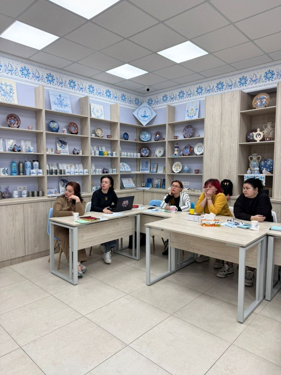 П’ятеро жінок сидять за столами під час робочої зустрічі або навчального заходу. Вони уважно слухають, на столах лежать ноутбук та робочі матеріали. Кімната оформлена стелажами з виставкою розписного посуду, кераміки та сувенірів у народному стилі.
