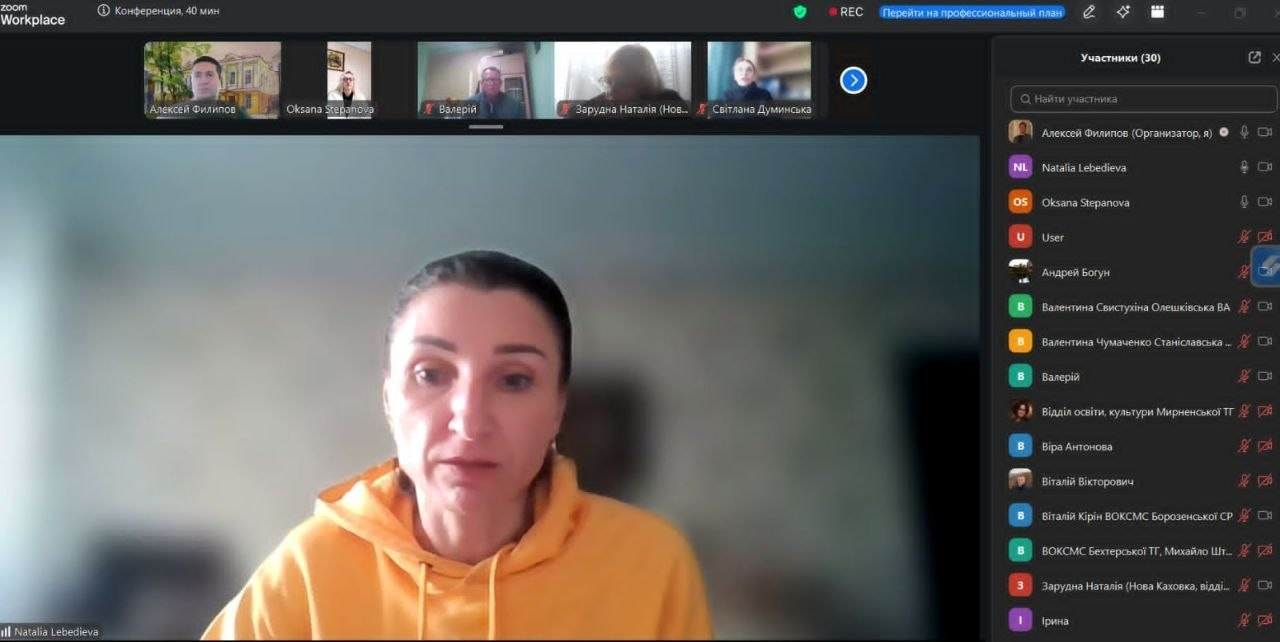 Скриншот вікна онлайн-конференції, на якому в центрі зображена жінка у жовтому худі під час виступу. У верхній частині екрана розташована панель із мініатюрами інших учасників, а праворуч — вертикальна колонка зі списком присутніх на зустрічі.