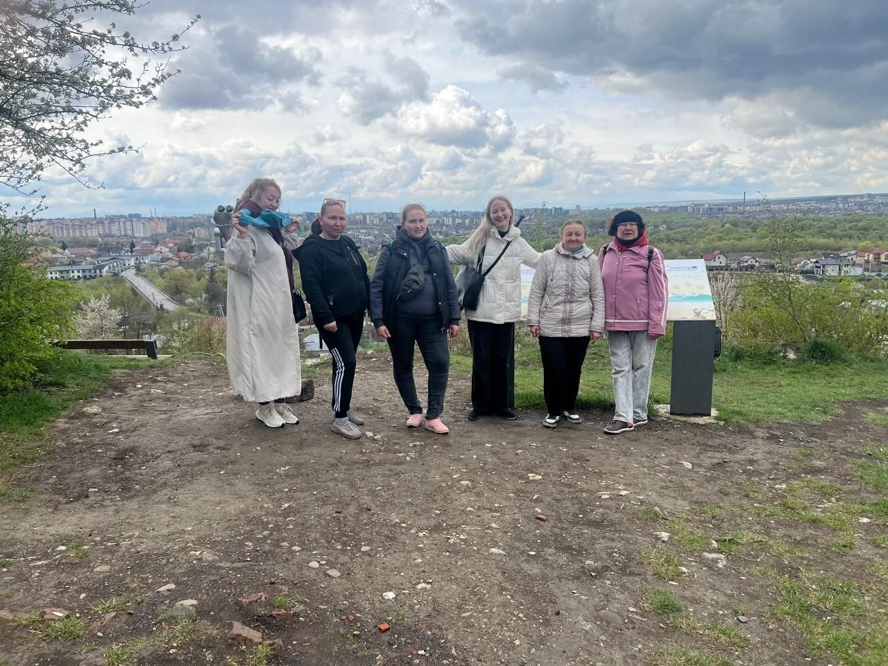 Група з шести жінок позує для фото на оглядовому майданчику на пагорбі. Позаду них відкривається панорама міста з житловими будинками та мостом під хмарним небом.