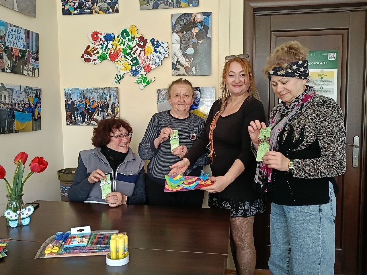 Чотири жінки показують виготовлені власноруч патріотичні вироби під час творчого заняття. На столі перед ними лежать матеріали для творчості, а стіна позаду прикрашена світлинами та кольоровою мапою України.