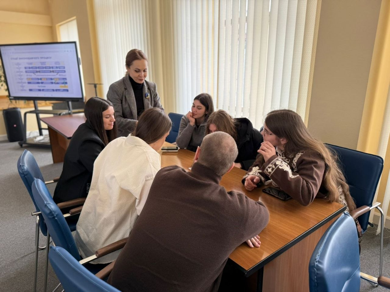 Група молодих людей сидить за столом у конференц-залі та активно обговорює проєкт під керівництвом жінки-модератора. На задньому плані видно великий екран із презентаційною схемою, приміщення світле з великими вікнами та жалюзі.