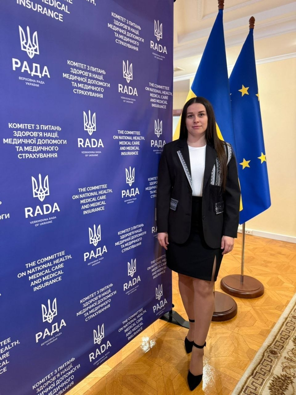Жінка в офіційному вбранні з елементами української вишивки позує на фоні банера Комітету Верховної Ради України з питань здоров'я нації. Поруч розташовані державний прапор України та прапор Європейського Союзу.