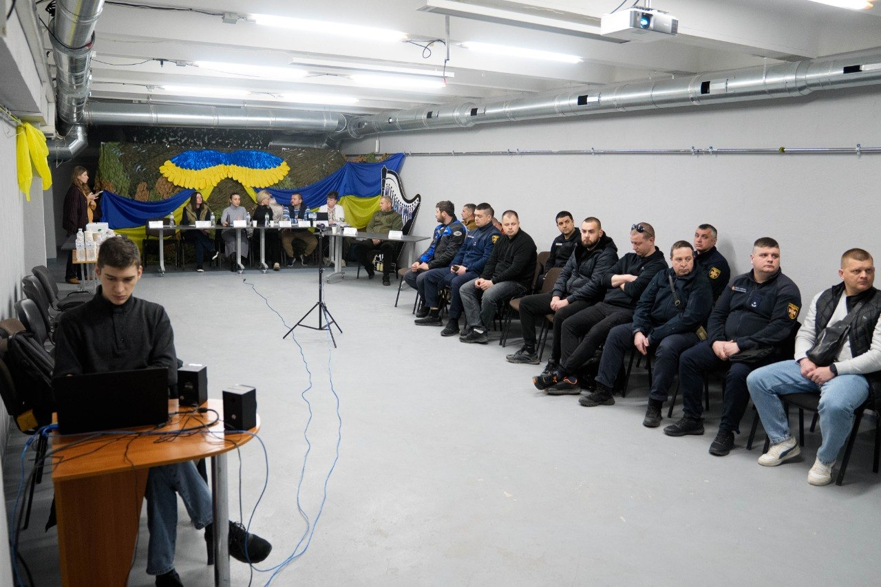 Група чоловіків у формі та цивільному одязі сидить у світлому приміщенні (схожому на облаштоване укриття) під час офіційного заходу. На задньому плані за столом сидять спікери на фоні стіни, прикрашеної державними прапорами України. На передньому плані ліворуч хлопець працює за ноутбуком.