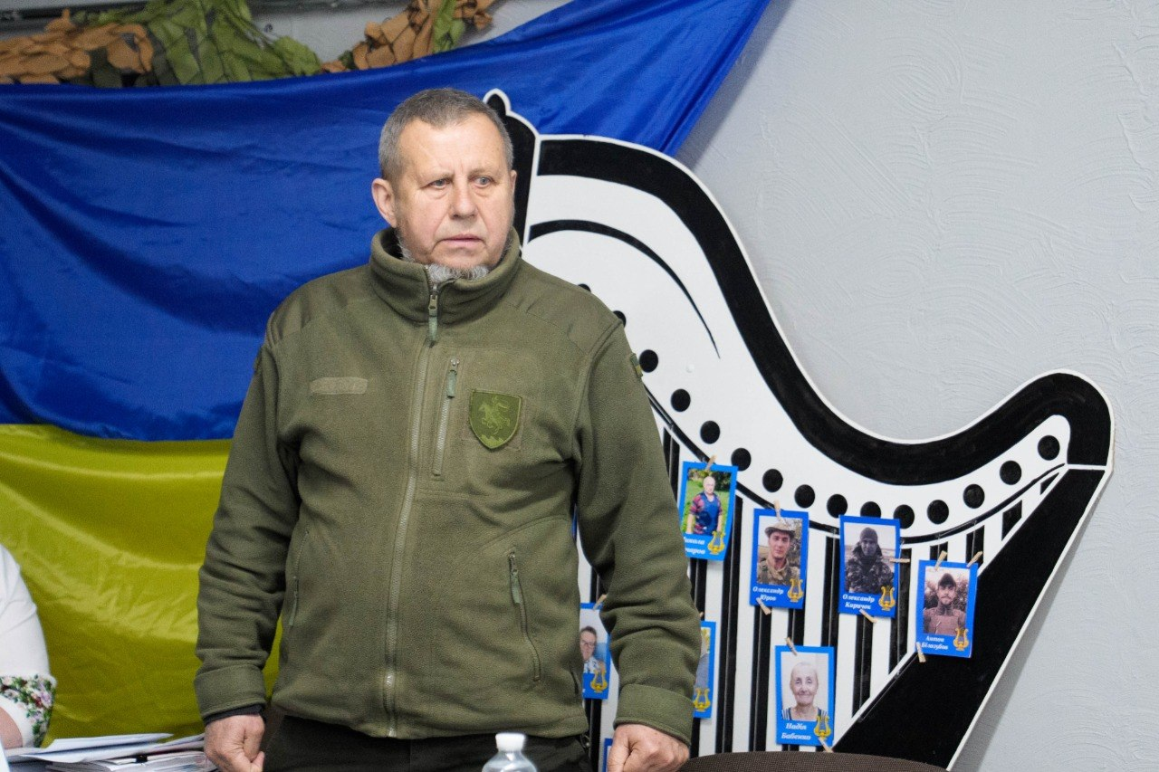 Чоловік у флісовій куртці військового зразка виступає на фоні державного прапора України. Поруч розташована декоративна конструкція у формі арфи, прикрашена невеликими фотографіями людей.