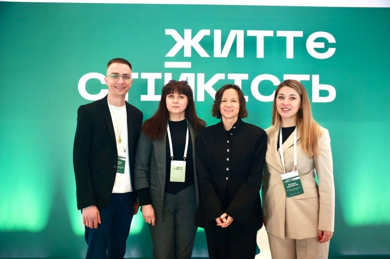 Група з чотирьох осіб (чоловік та три жінки) позує для фото на фоні зеленого банера з великим білим написом «ЖИТТЄСТІЙКІСТЬ». Учасники заходу одягнені в офіційно-діловому стилі та мають на шиї бейджі.