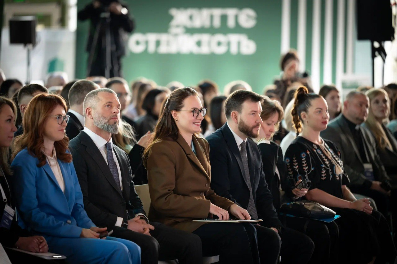 Учасники форуму сидять у залі та уважно слухають виступ. Люди одягнені в офіційно-діловому та етностилі. На задньому плані видно зелений банер із написом «ЖИТТЄСТІЙКІСТЬ», професійну відеокамеру на штативі та звукове обладнання.